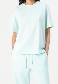 Miętowo-zielona, oversize'owa koszulka wykonana z miękkiego materiału, z krótkimi rękawami i małym białym logo na lewej stronie klatki piersiowej.