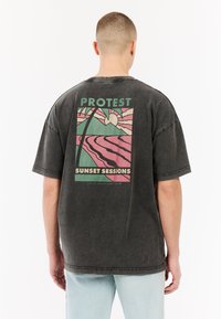 Over-sized zwart katoen t-shirt met een kleurrijke grafische print op de achterkant met palmbomen, zon en de tekst "PROTEST SUNSET SESSIONS."