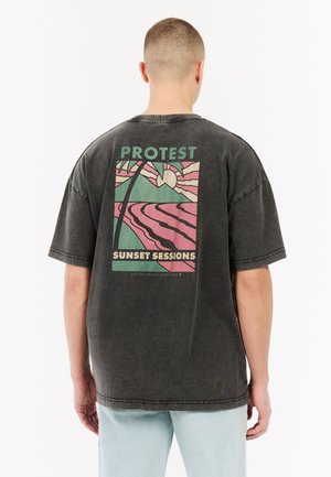 Oversized black cotton t-shirt featuring a colorful graphic print on the back with palm trees, sun, and the text "PROTEST SUNSET SESSIONS."