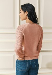 Gaiši rozā džemperis ar mežģīņu raksta piedurknēm, ribots apakšmala un apaļa kakla izgriezums, saskaņots ar zilajām džinsa biksēm. Mīksts, fakturēts audums.