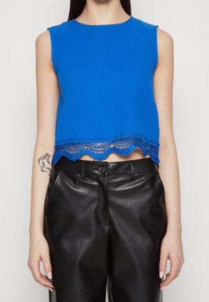 Kvinde iført en ærmeløs, lys blå cropped top med blonderkant, kombineret med højttaljede sorte læderbukser, der delvist viser en tatovering på venstre arm.