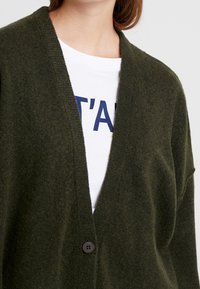 Dark green, textured cardigan with a V-neck design, single button closure, and loose fit, layered over a white T-shirt with navy text.