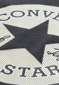 Juodo žvaigždės logotipas ant tekstūruoto kreminio fono su įdubimais, kuriame rašomas žodis „CONVERSE“ storomis juodomis raidėmis ir yra parašo detalė.