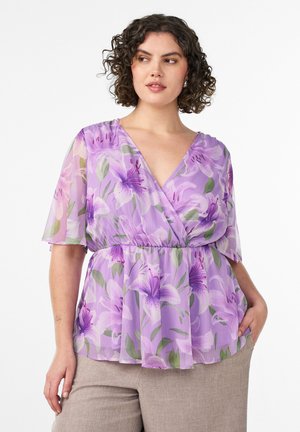 Mujer con cabello rizado que lleva una blusa cruzada morada con estampado floral y mangas cortas, y pantalones beige, posando con una mano en el bolsillo.