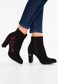 Bottines en daim noir avec broderie florale rouge et marron, dotées d'une fermeture éclair et d'un talon bloc orné d'un accent floral.