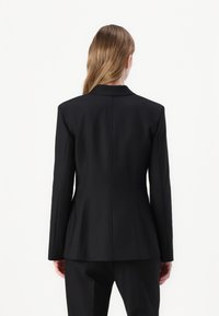 Černý přizpůsobený blazer se strukturovanými ramenními výztuhami, klopovým límcem a jemným detailem v zadním švu. Hladká tkanina s elegantním, úzkým střihem.