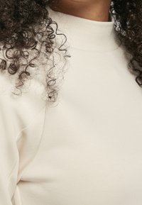 Sudadera beige con cuello alto y textura suave, de diseño ajustado y costuras ligeramente curvas para un aspecto entallado.