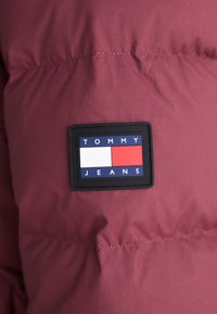 Maroon puffer jacket section with a black rectangular patch featuring the text "TOMMY" and "JEANS" in white and blue, with red and white accents.