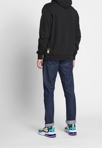 Black hoodie with kangaroo pocket, dark blue jeans, and multicolored sneakers. Notable details: yellow accent on hoodie hem and sneaker design.
