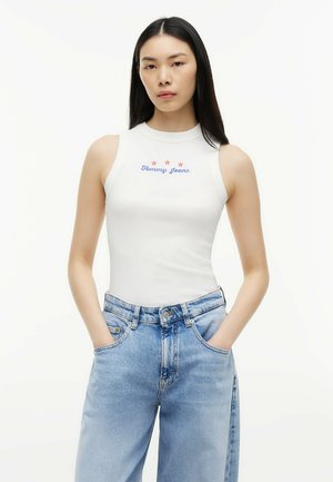 Młoda kobieta z długimi czarnymi włosami, ubrana w biały bezrękawnik "Tommy Jeans" i jasnoniebieskie, luźne, wysokie jeansy z rękami w kieszeniach.