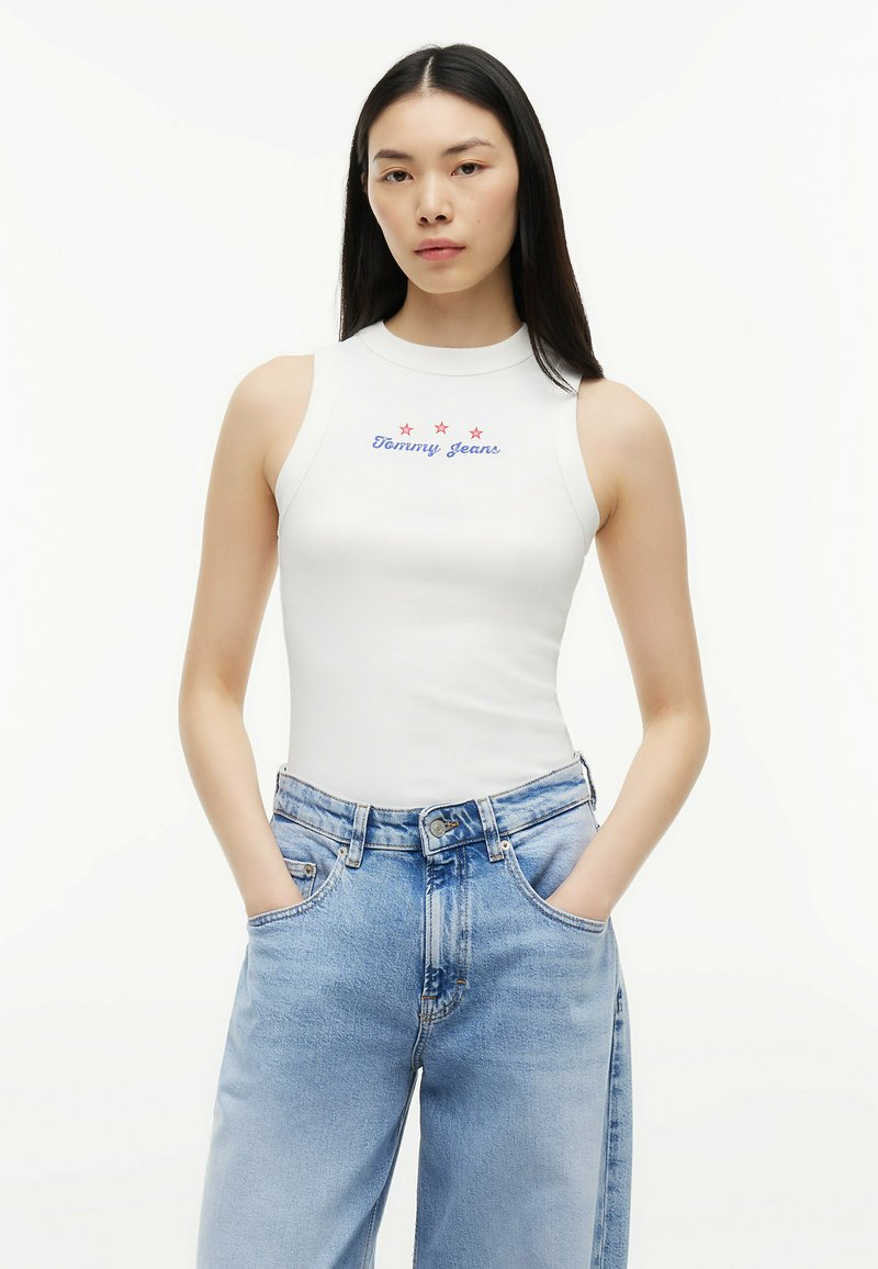 Jeune femme aux longs cheveux noirs portant un haut blanc sans manches "Tommy Jeans" et un jean large taille haute bleu clair, les mains dans les poches.