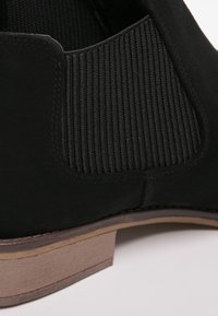 Czarny zamszowy botek na kostkę z elastycznymi bokami i drewnianym, warstwowym obcasem, prezentuj�ący gładką teksturę i minimalistyczny design.