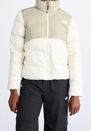 Kremowa beżowa pikowana kurtka z frontowym zamkiem błyskawicznym noszona z czarnymi spodniami, z logo The North Face na kurtce i spodniach.