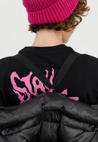 Svart boblejakke med blank tekstur og minimal søm, holdt av svarte stropper. Svart t-skjorte med rosa "STAY" grafikk på baksiden. Rosa strikket lue.