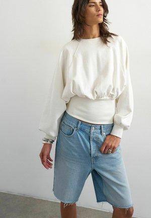 Mulher a usar uma camisa branca de mangas compridas e larga e calções de ganga azuis claros até ao joelho com bainhas desfiadas, a posar junto a uma parede simples.