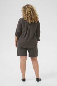 Femme aux cheveux blonds bouclés, portant un haut à manches longues sombre, un short assorti et des ballerines noires, debout dos à nous.