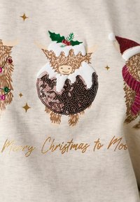 Vache des Highlands festive portant un costume de pudding de Noël avec houx et glaçage, posée sur un tissu crème avec texte doré et étoiles.