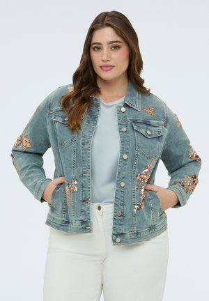 Žena s dlouhými hnědými vlasy, oblečená v světle modré džínové bundě s růžově zlatými květinovými flitry a bílých kalhotách, ruce v kapsách bundy.