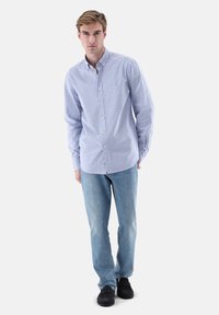 Langärmeliges Hemd mit Knöpfen in hellblauen und weißen vertikalen Streifen, kombiniert mit hellblauen Jeans und schwarzen Schuhen. Einfaches Taschen-Detail.