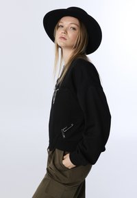 Zwarte sweatshirt met een ontspannen pasvorm, ritszakken en een gestructureerd oppervlak. Gecombineerd met olijfgroene broek en een brede zwarte hoed.