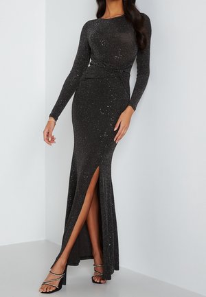 Vestido largo ajustado de manga larga negro con purpurina, con abertura frontal en el muslo, combinado con sandalias negras de tacón alto con tiras adornadas con piedras brillantes.