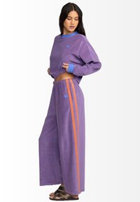 Conjunto de loungewear a rayas moradas que incluye una camiseta corta de corte suelto y pantalones de pierna ancha con franjas laterales naranjas. Tejido suave y texturizado.
