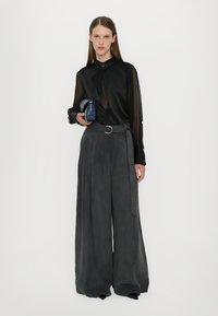 Blusa nera trasparente con chiusura a bottoni, abbinata a pantaloni grigi a gamba larga con cintura e dettagli piegati. Borsa blu in mano.
