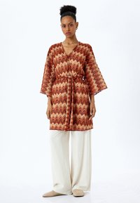 Röd och orange zigzag-mönstrad kimono med snörliv, vida ärmar, bärs över krämfärgad vida byxor och beige tofflor.