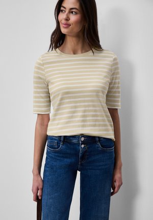 Mujer con camiseta de manga corta a rayas beige y blanca y jeans azules, de pie frente a un fondo liso gris claro.