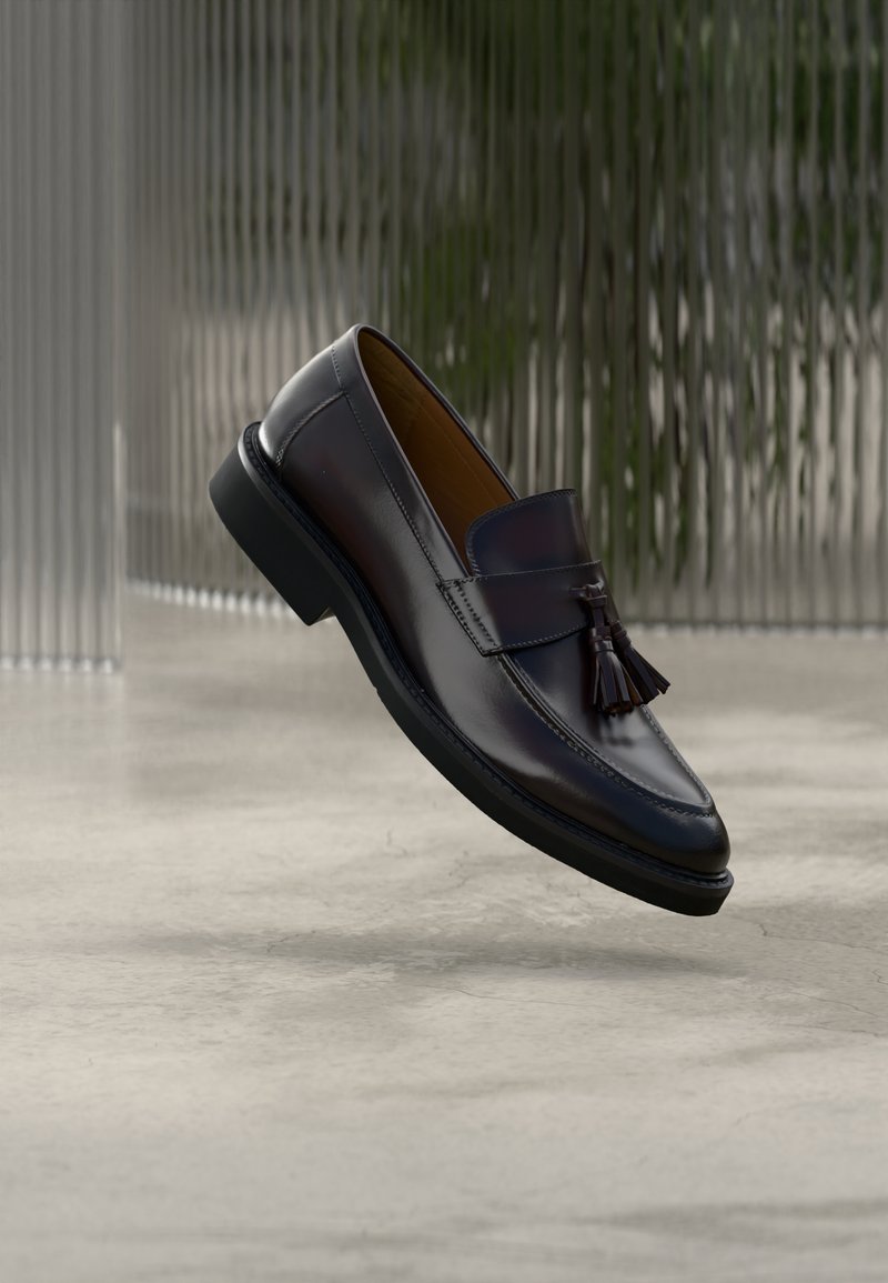 Mocassins en cuir brun foncé à finition brillante, ornés d'un pompon décoratif et d'un bout arrondi, posés sur une surface en béton.