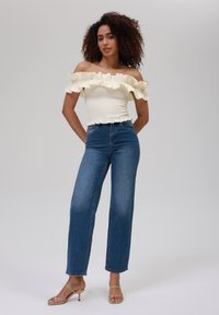Top blanco off-the-shoulder con ribetes de volantes, combinado con jeans azules de tiro alto. La modelo lleva tacones de tiras en tono nude. Textura simple y suave.