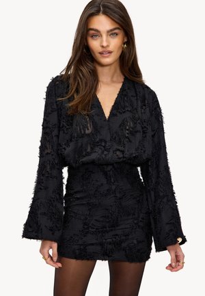 Mujer con un vestido mini negro de manga larga y textura, con mangas amplias y escote en V, combinado con medias negras transparentes y pendientes de aro dorados.