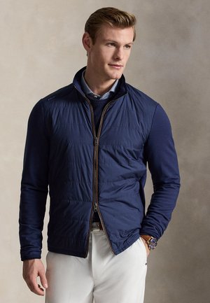 Moški v temno modri prešiti jakni z zadrgo čez modrim puloverjem in svetlo modro srajco s ovratnikom, oblečen v bele hlače in z uro na levi zapestju.