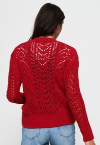 Pull en tricot rouge avec un motif ajouré, des poignets et un ourlet côtelés, présentant une silhouette décontractée. L'arrière-plan est blanc uni.