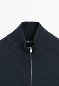 Pull bleu marine avec un col montant et une fermeture éclair argentée. Présente une texture côtelée au niveau du col et des coutures des épaules. Étiquette de la marque visible à l'intérieur.