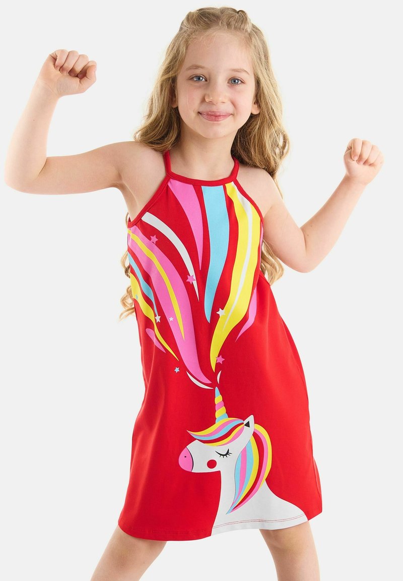 Niña pequeña sonriendo y flexionando los brazos, vestida con un vestido rojo con un diseño colorido de unicornio y arcoíris sobre un fondo blanco.