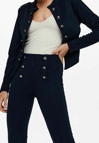 Veste bleu marine cintrée avec fermeture à boutons, associée à un pantalon taille haute avec boutons métalliques croisés et coupe slim.