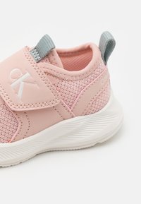 Różowy but sportowy z gładką syntetyczną cholewką, siateczkowymi wstawkami, regulowanym paskiem i teksturowaną białą podeszwą. Szara pętelka do ściągania z tyłu.