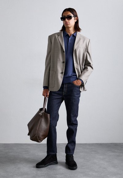 Homme portant un blazer gris, un polo bleu, un jean sombre, des baskets noires, des lunettes de soleil, tenant un grand sac en cuir brun dans un espace intérieur neutre.