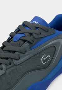 Αθλητικά παπούτσια Lacoste με γκρι επάνω μέρος από δίχτυ και δέρμα, μπλε λεπτομέρειες, υφασμένα κορδόνια και ασημένιο λογότυπο στο πλάι. Υφή στη σόλα από καουτσούκ.