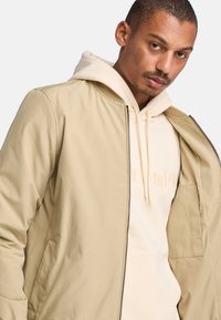 Chaqueta bomber beige con una textura suave y mate y cuello acanalado, superpuesta a una sudadera ligera beige con un logo. Con cremallera en la parte delantera.