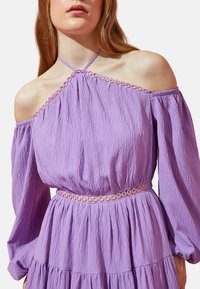 Lila schulterfreies Kleid mit langen Ärmeln, gewellter Textur, elastischem Taillenbund und kreisförmigen Verzierungen entlang des Ausschnitts und der Taille.