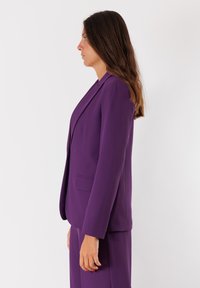 Blazer viola con vestibilità su misura, revers e tasca singola. Tessuto liscio con design strutturato. Abbinato a pantaloni coordinati.