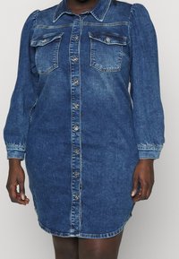 Robe en denim boutonnée avec manches longues bouffantes, deux poches poitrine à rabat et coutures contrastantes sur une personne à la peau foncée.