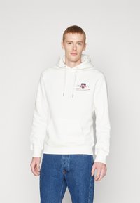 Sweatshirt à capuche blanc avec poche avant, arborant un logo rouge foncé et bleu. Accompagné d'un jean en denim bleu. Design texturé et décontracté.