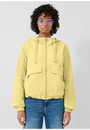 Frau mit lockigem Haar, die übergroße Brille mit blauen Gläsern, eine gelbe Kapuzenjacke mit Reißverschluss und blaue Jeans trägt, steht vor einem einfarbigen Hintergrund.