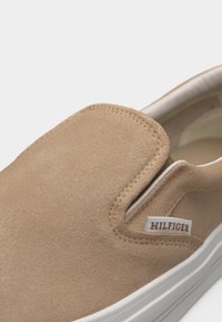 Zapatilla sin cordones beige con suela blanca y pequeña etiqueta de tela "Hilfiger" en el lateral, que presenta una textura suave similar al ante y paneles laterales elásticos.