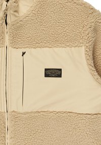 Gilet in fleece beige con un pannello cremoso e liscio, chiusura frontale a zip e un'etichetta con logo ricamato. Presenta una texture morbida e cuciture a contrasto.