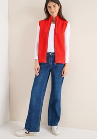 Rode mouwloze geribde spencer over een wit shirt met lange mouwen, gecombineerd met wijde blauwe jeans en witte sneakers met een bordeauxrood accent.