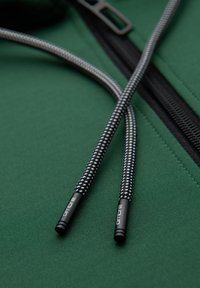 Cordons de sweat à capuche à motif noir et blanc avec embouts métalliques reposant sur un tissu vert près d'une fermeture éclair noire partiellement ouverte.